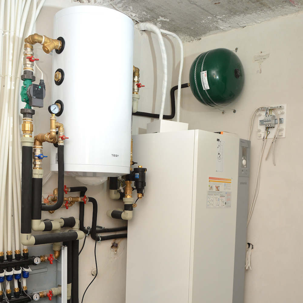White vertical boiler unit with copper pipes, gauges, and a green expansion tank in a utility room.