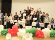 Group of young award winners on stage holding certificates and tote bags, wearing medals, with colorful balloons in the foreground and a 1st place podium nearby.