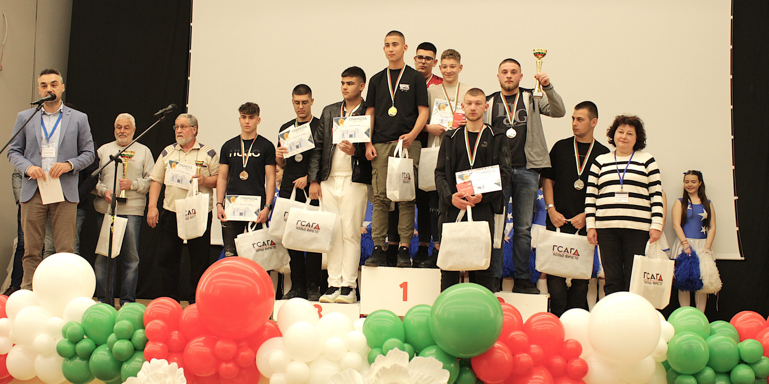Group of young award winners on stage holding certificates and tote bags, wearing medals, with colorful balloons in the foreground and a 1st place podium nearby.