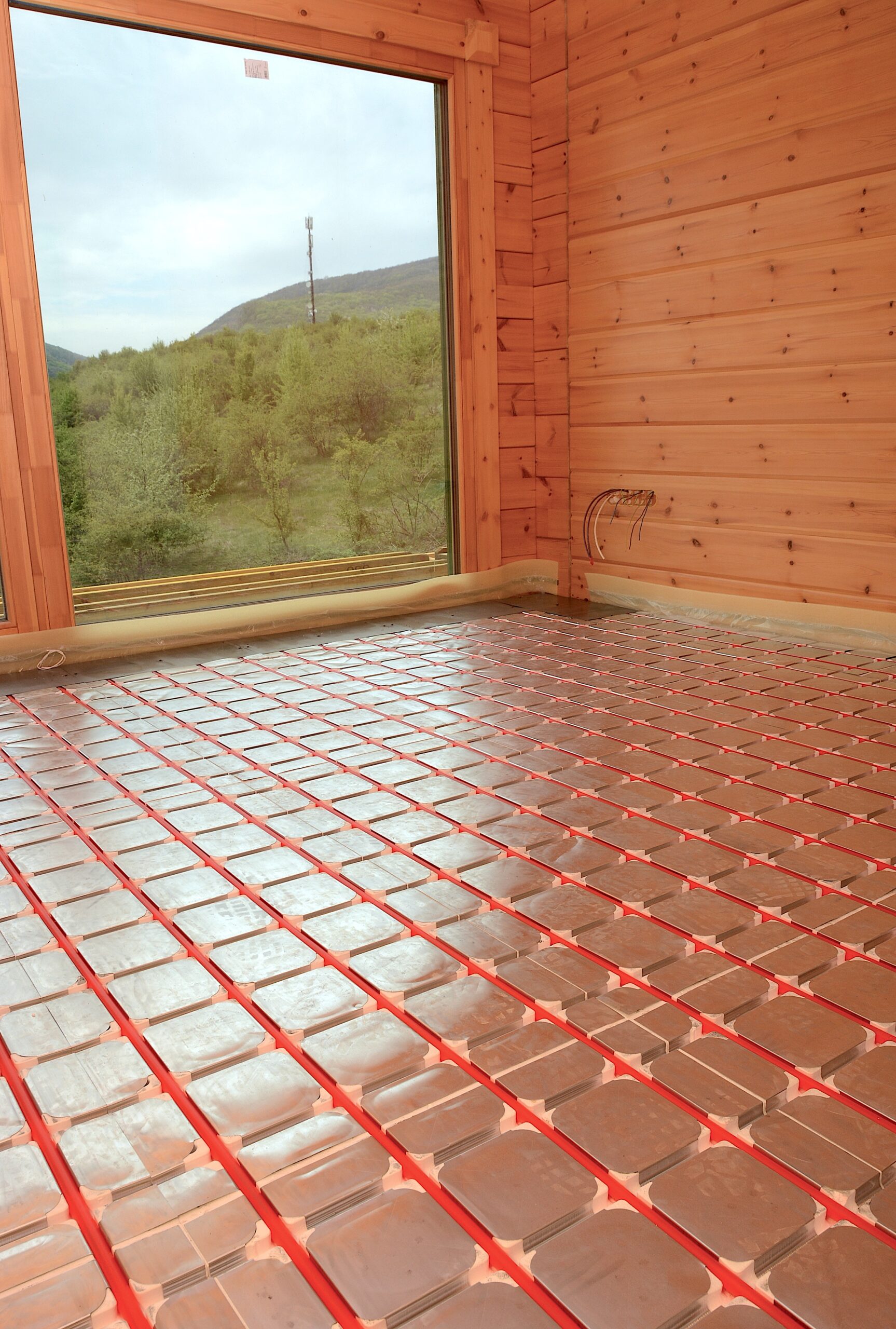 Interior of a wooden cabin under construction, showing radiant floor heating mats and wiring beneath a large window with a forested landscape outside