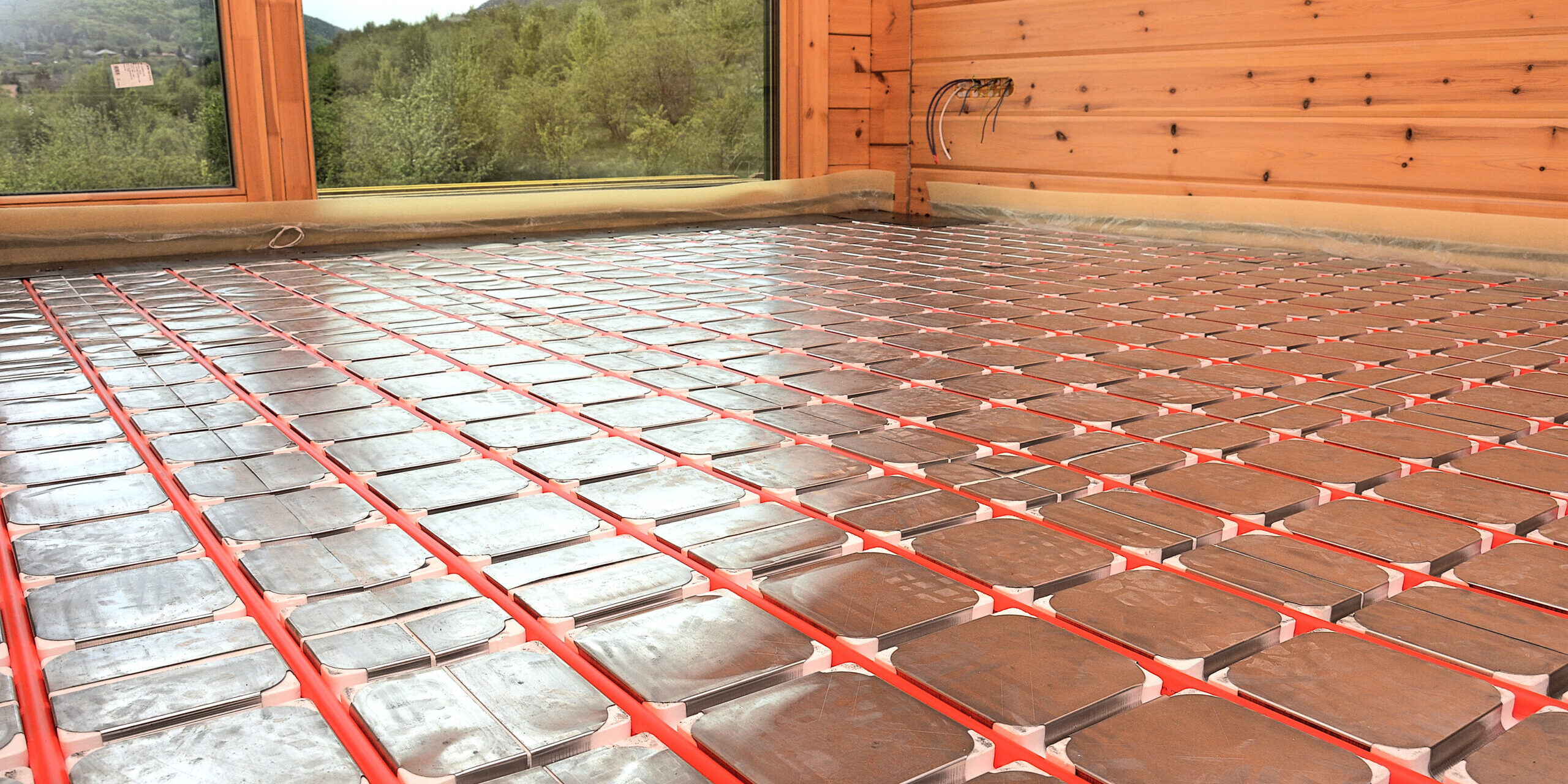 DSC_2617 Underfloor radiant heating mats installed in a wood-paneled room, red pipes laid between metal panels. View shows forested hills through the window.