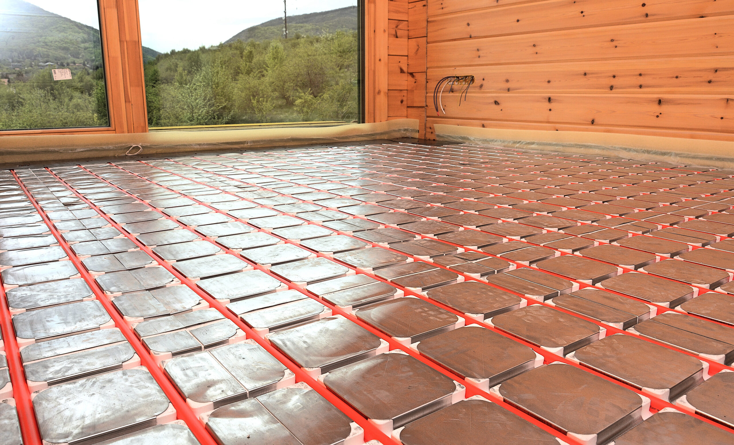 Underfloor radiant heating mats installed in a wood-paneled room, red pipes laid between metal panels. View shows forested hills through the window.