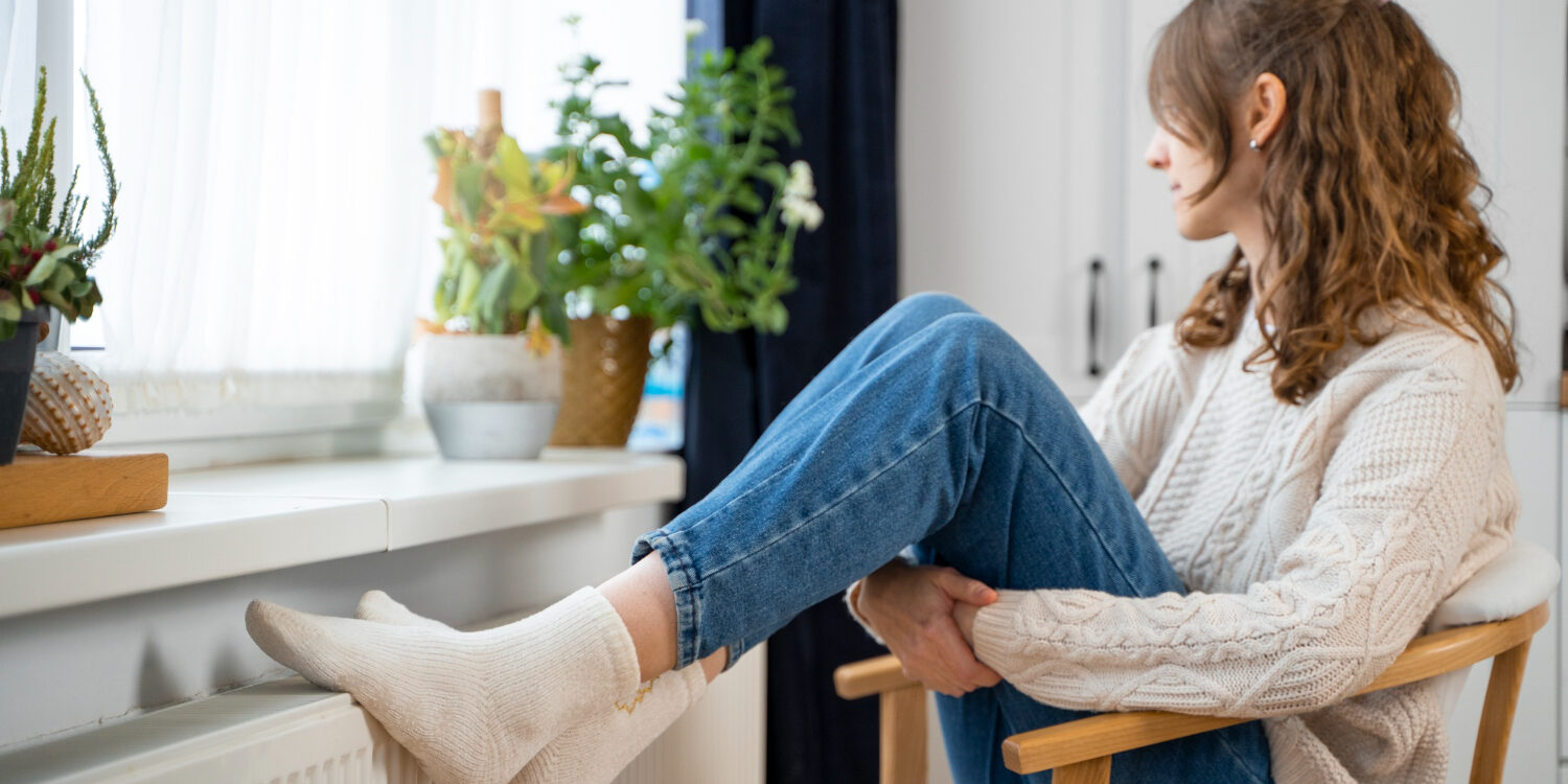 Woman relaxing indoors, sitting in a chair with feet up on a radiator by a sunny window with potted plants, wearing a cream knit sweater and jeans.
