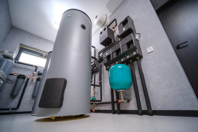 Industrial mechanical room with a tall gray cylindrical tank, teal expansion tank, and exposed piping.