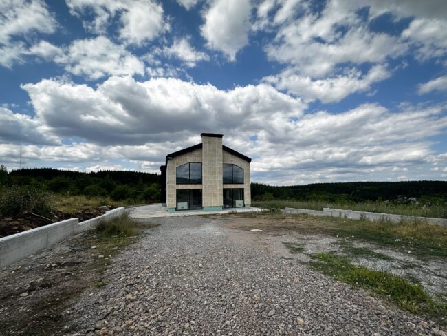 Modern, two-story house with large windows sits on a gravel lot under a blue sky filled with white clouds, in a rural construction site.